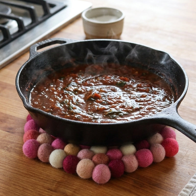 Black cast iron skillet with simmering food on a wooden countertop, surrounded by colorful pom-pom trivets.