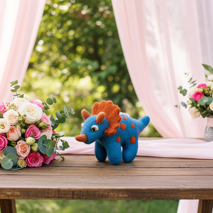 Blue wool felt triceratops hanging decoration on a wooden table with flowers and curtains in the background.