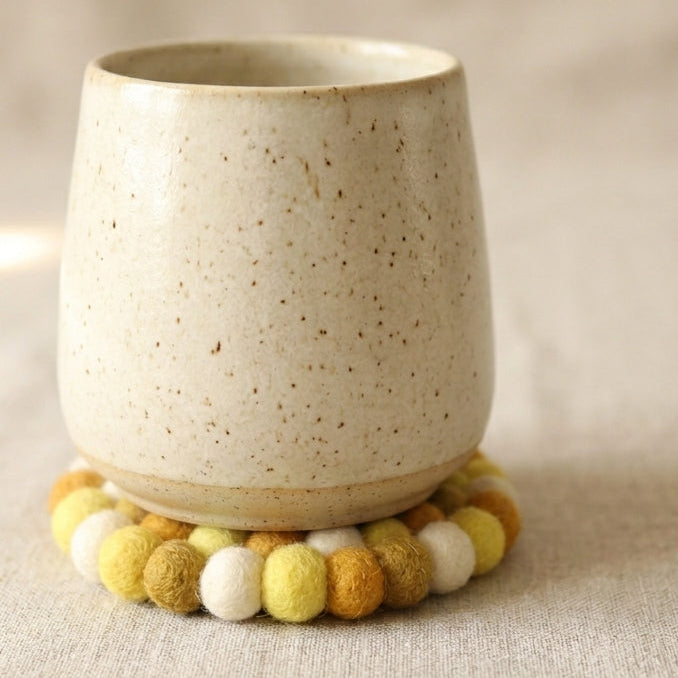 Ceramic cup with a textured surface on  Mustard Yellow wool felt coasters handmade in Nepal, warm absorbent non-scratch drink coasters for wooden tables, teacher gifts, host gifts and new home buyers ON a beige background