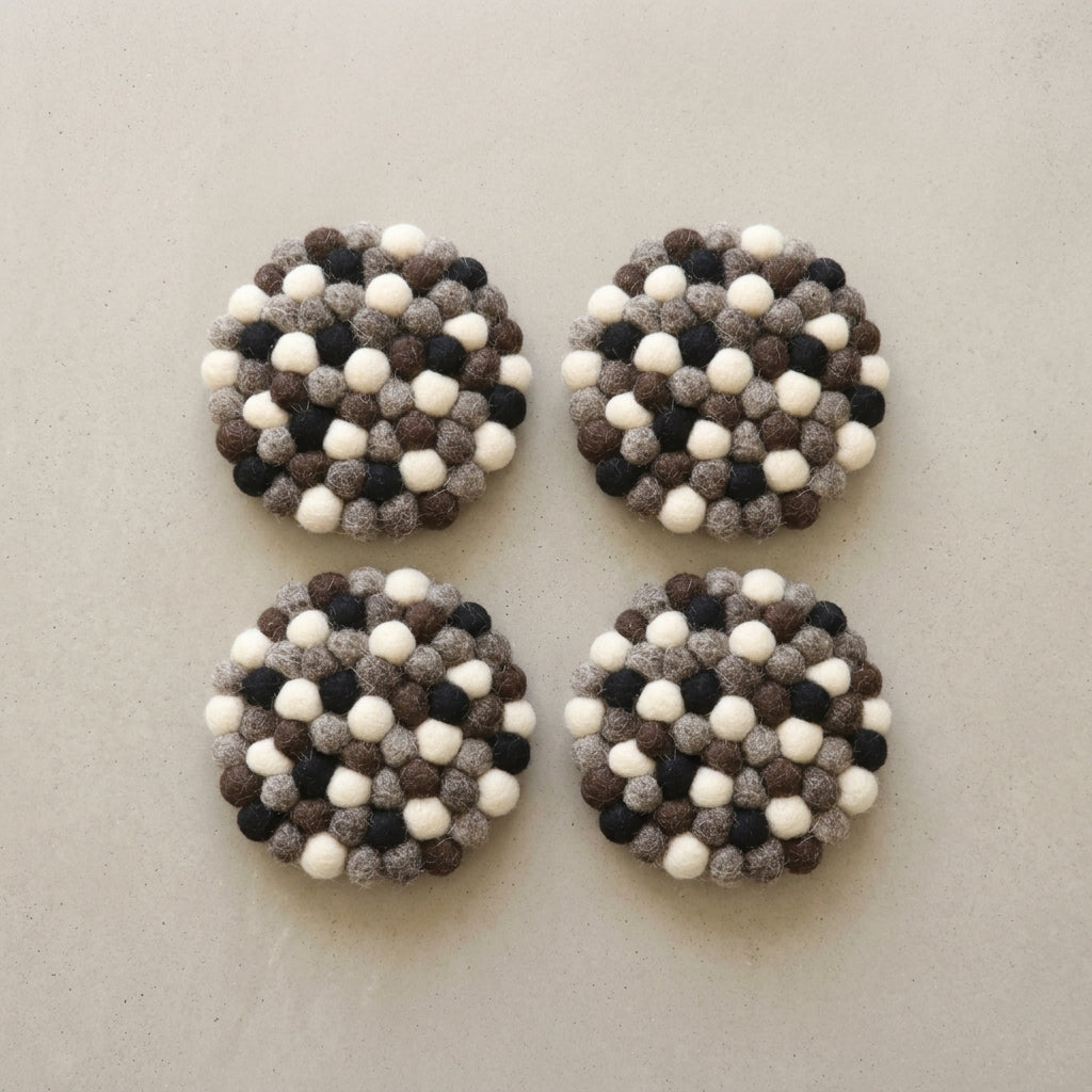 Four round Monochrome Pebble Black wool felt coasters handmade in Nepal, neutral absorbent non-scratch drink coasters for wooden tables, minimalist homes, host gifts and new home buyers with a pattern of white, black, and gray balls on a light gray background.
