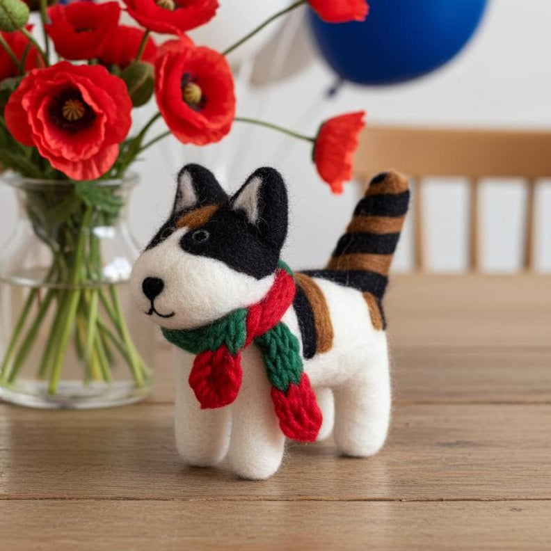 Handmade wool felt black and white cat ornament with hanging thread next to a vase of red flowers and balloons on a wooden table.