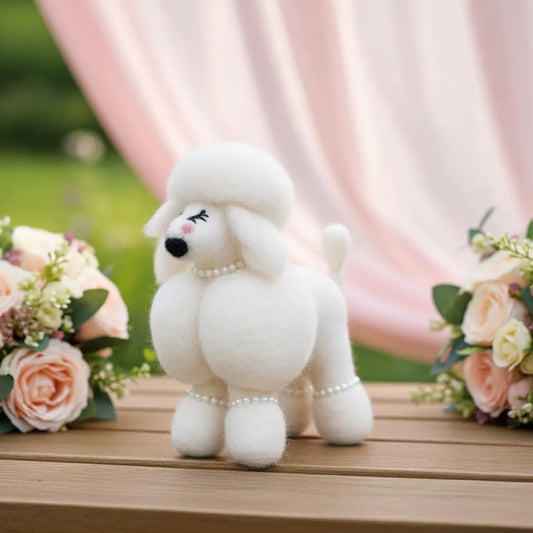 Handmade wool felt poodle dog ornament  with a pearl necklace on a wooden surface with flowers and pink curtains in the background.