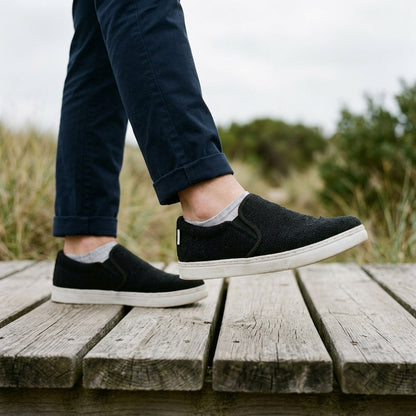 Person wearing black pure hemp vegan slip-on shoes with white rubber soles on a wooden platform outdoors.
