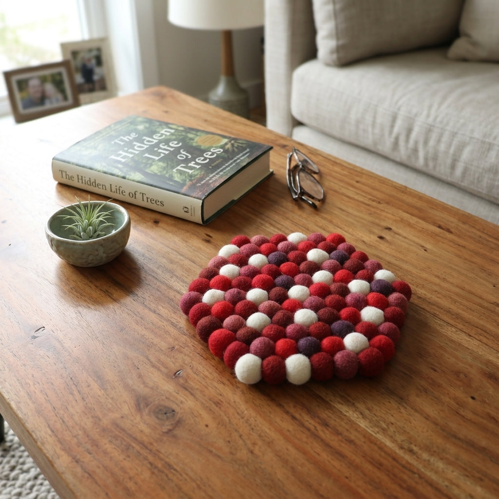 Decorative felt ball coaster on a wooden coffee table with a book and small plant.