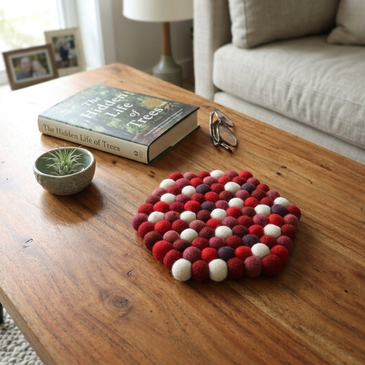 Decorative felt ball coaster on a wooden coffee table with a book and small plant.