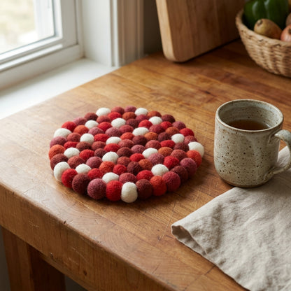 Round felt trivet with red and white pattern on a wooden surface next to a mug.