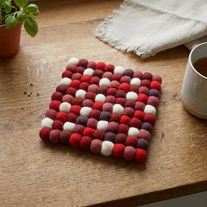 Woven trivet with red, white, and purple pattern on a wooden surface with a mug and plant.