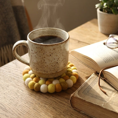 Steaming cup of coffee on a tMustard Yellow wool felt coasters handmade in Nepal, warm absorbent non-scratch drink coasters for wooden tables, teacher gifts, host gifts and new home buyers with an open book beside it on a wooden surface.