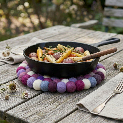 Black frying pan with vegetables on a colorful felt trivet on a wooden table outdoors.