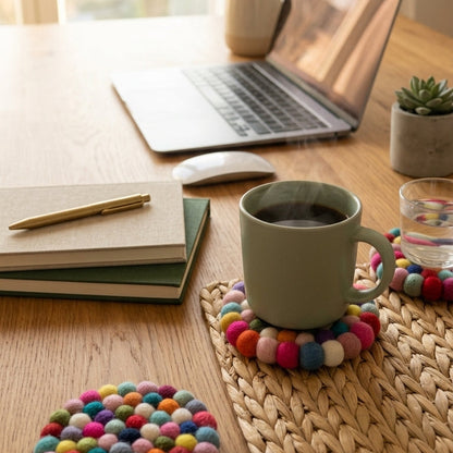 Desk setup with laptop, mug, and Confetti Mix multi wool felt coasters handmade in Nepal, colourful absorbent non-scratch drink coasters for wooden tables, host gifts, housewarming gifts and stocking fillers on a wooden surface.