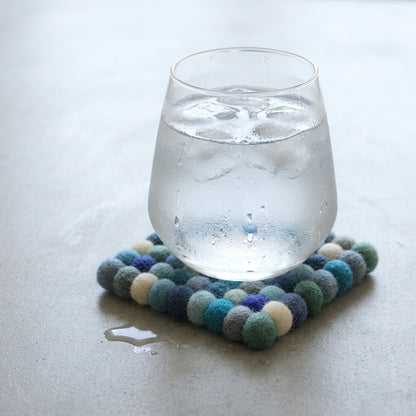 Glass of water on aOcean Mix Blue wool felt coasters handmade in Nepal, coastal absorbent non-scratch drink coasters for wooden tables, coffee lover gifts, host gifts and new home buyerswith a light gray background