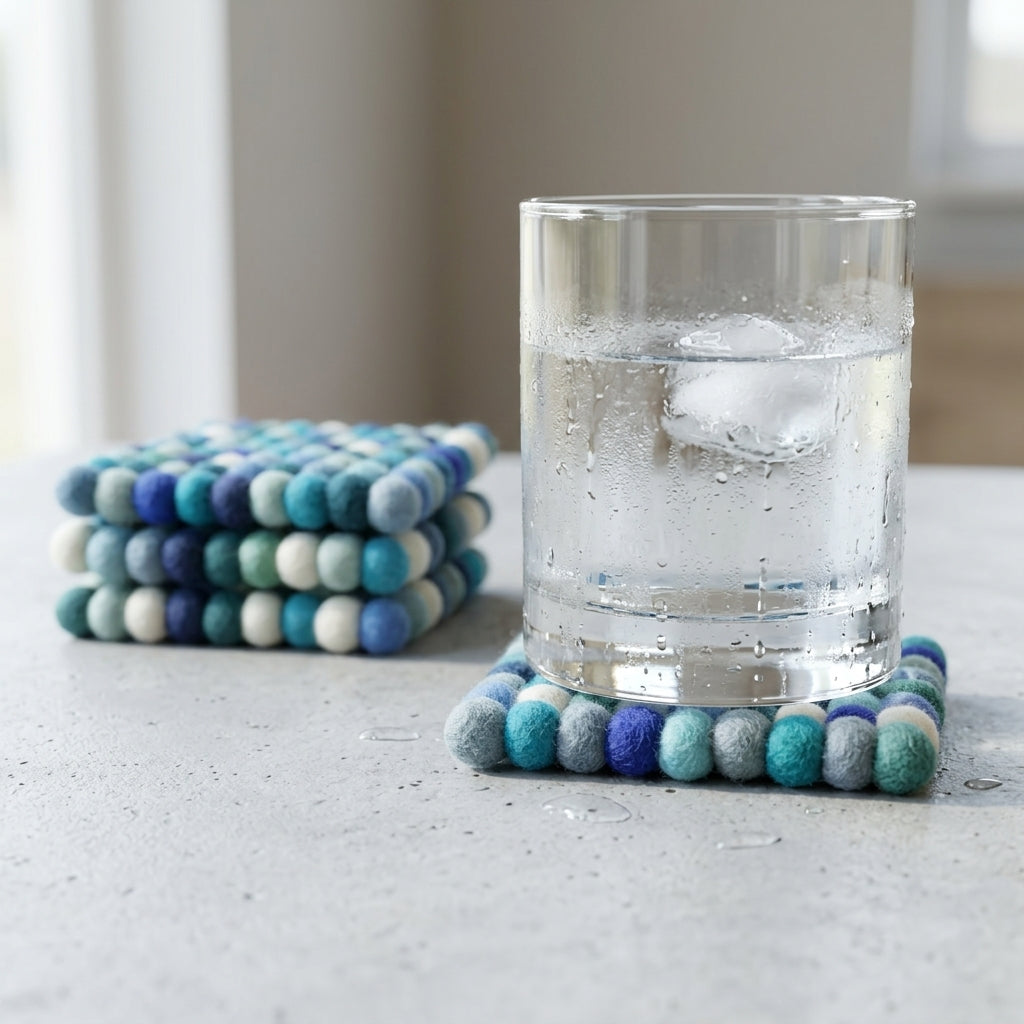 Stack ofOcean Mix Blue wool felt coasters handmade in Nepal, coastal absorbent non-scratch drink coasters for wooden tables, coffee lover gifts, host gifts and new home buyers with a glass of water on a light surface.