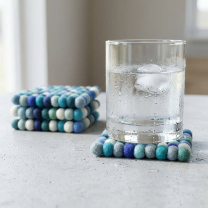 Stack ofOcean Mix Blue wool felt coasters handmade in Nepal, coastal absorbent non-scratch drink coasters for wooden tables, coffee lover gifts, host gifts and new home buyers with a glass of water on a light surface.