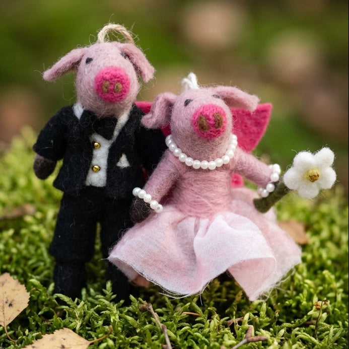 Handmade pig couple felt ornament for wedding or anniversary gift, one in a suit and the other in a dress, standing on green moss with a blurred natural background.
