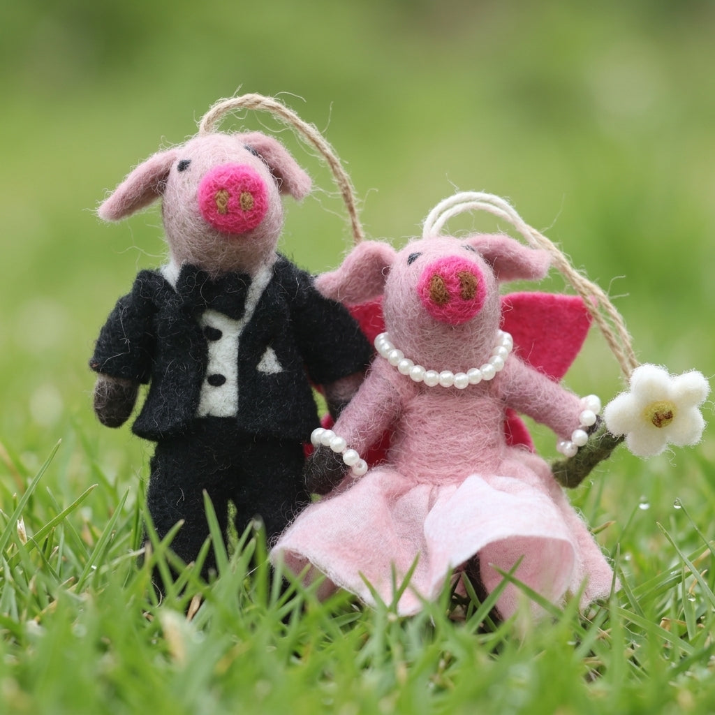 Bride and groom pig ornament made from wool felt, one in a tuxedo and the other in a dress with wings, standing on grass.