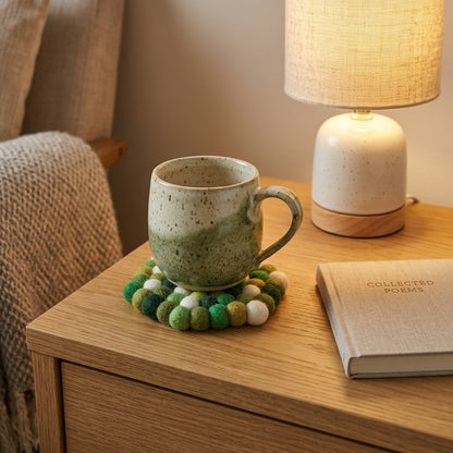 Ceramic mug onForest Mix Green wool felt coasters handmade in Nepal, botanical absorbent non-scratch drink coasters for wooden tables, plant lover gifts, host gifts and new home buyers with a lamp and book on a wooden surface
