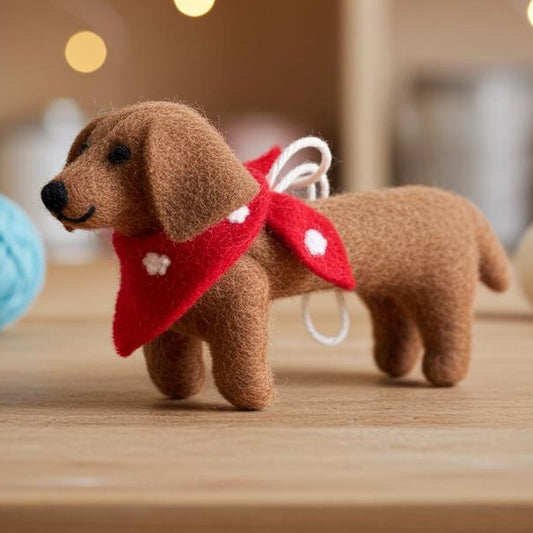 Handmade wool felt dachshund dog ornament  with a red scarf on a wooden surface with blurred lights in the background