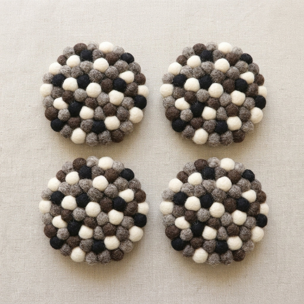Four round Monochrome Pebble Black wool felt coasters handmade in Nepal, neutral absorbent non-scratch drink coasters for wooden tables, minimalist homes, host gifts and new home buyerswith a pattern of white, black, and brown balls on a beige fabric background.