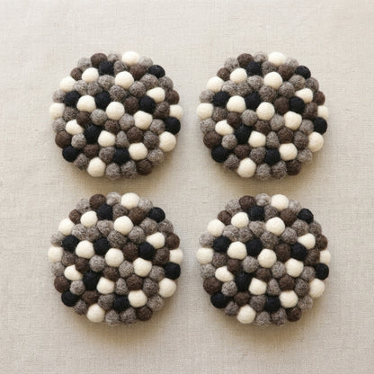 Four round Monochrome Pebble Black wool felt coasters handmade in Nepal, neutral absorbent non-scratch drink coasters for wooden tables, minimalist homes, host gifts and new home buyerswith a pattern of white, black, and brown balls on a beige fabric background.