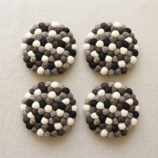 Four round Monochrome Pebble Black wool felt coasters handmade in Nepal, neutral absorbent non-scratch drink coasters for wooden tables, minimalist homes, host gifts and new home buyerswith a pattern of white, black, and brown balls on a beige fabric background.