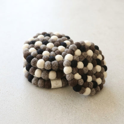 Two Monochrome Pebble Black wool felt coasters handmade in Nepal, neutral absorbent non-scratch drink coasters for wooden tables, minimalist homes, host gifts and new home buyers with a pattern of brown, black, and white on a light background.