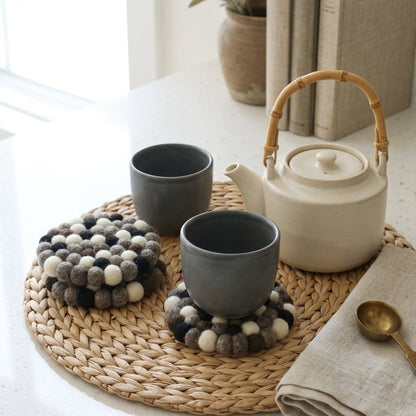 Tea set with two gray cups, a white teapot, and Monochrome Pebble Black wool felt coasters handmade in Nepal, neutral absorbent non-scratch drink coasters for wooden tables, minimalist homes, host gifts and new home buyers on a light surface.