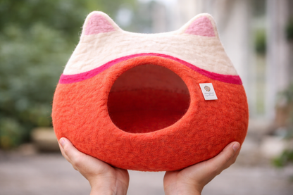 Cat-shaped pet bed with red body and white face held by a hand outdoors.