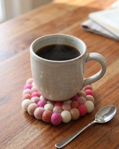 White mug with steaming coffee on a pink and white felt coaster on a wooden table.